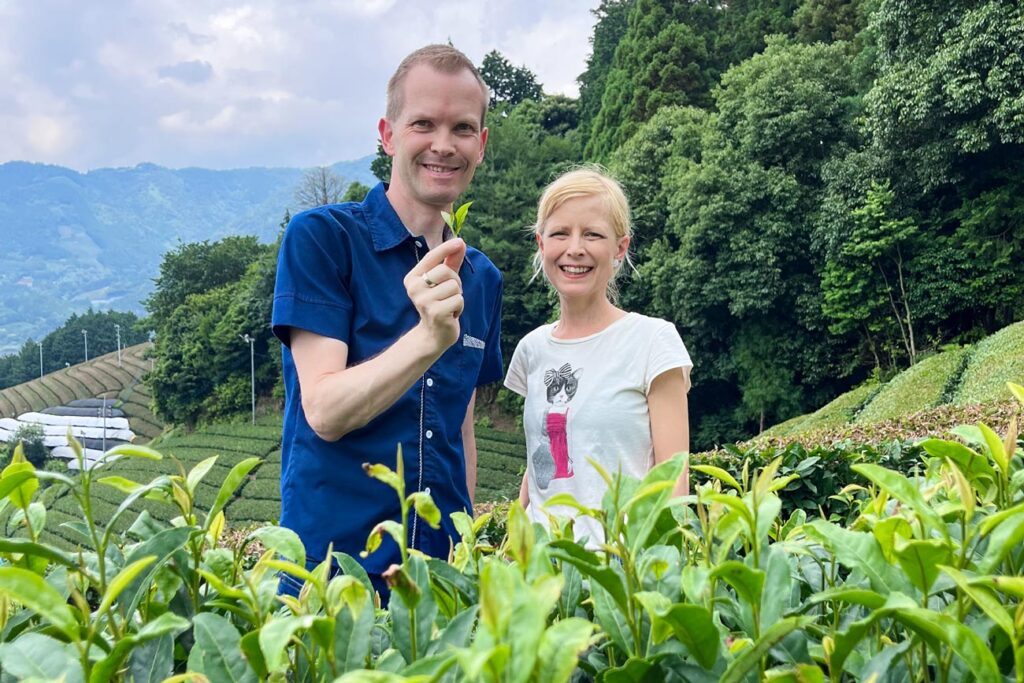 Holger Müller und Dr. Christin Müller-Wenzel stehen in einem grünen Teefeld und halten ein frisch gepflücktes Teeblatt, umgeben von Teeplantagen und bewaldeten Hügeln im Hintergrund.