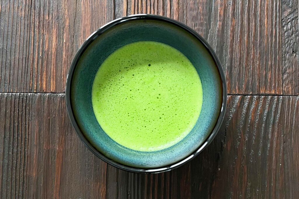 Schale mit frisch zubereitetem, hellgrünem Matcha-Tee auf dunklem Holztisch aus der Draufsicht.