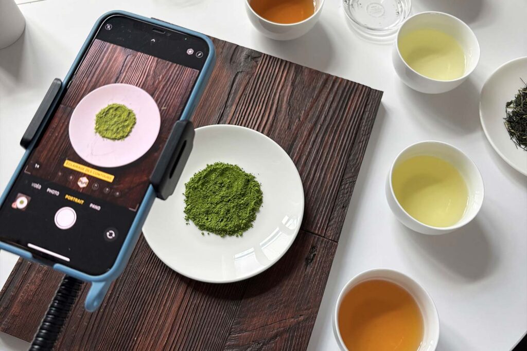 Smartphone fotografiert Matcha-Pulver auf einem Teller, umgeben von Teetassen und verschiedenen Teesorten auf einem Tisch.