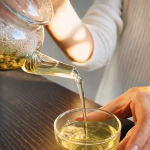 Einschänken von Tee aus einer Glaskanne in eine kleine Glasschale. Der Tee ist hellgelb.