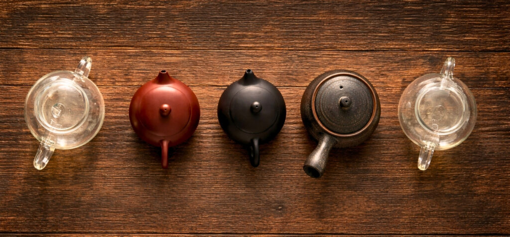 Draufsicht auf fünf Teekannen in einer horizontalen Reihe auf brauner Holzoberfläche: zwei Glaskannen außen, dazwischen eine rotbraune, eine schwarze und eine dunkel strukturierte Teekanne.