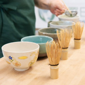 Mehrere Matcha-Schalen aus Keramik stehen auf einem hellen Holztisch, davor sind vier Bambus-Matchabesen (Chasen) aufgereiht. Im Hintergrund siebt eine Person in grüner Schürze Matcha-Pulver durch ein Sieb in eine Schale; rechts steht eine gläserne Teekanne.