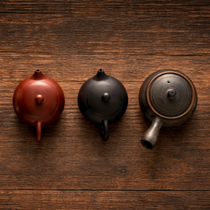 Draufsicht auf drei Teekannen in einer horizontalen Reihe auf brauner Holzoberfläche: eine rotbraune, eine schwarze und eine dunkel strukturierte Teekanne.