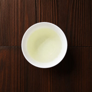 Tasse mit hellgrünem aufgebrühtem Gyokuro-Grüntee auf dunklem Holztisch aus der Draufsicht. Produkt aus dem iūgen Shop.