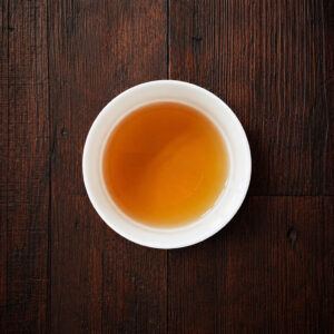 weiße Tasse mit bernsteinfarbenem aufgebrühtem Hōjicha-Tee auf dunklem Holztisch aus der Draufsicht. Produkt aus dem iūgen Shop.