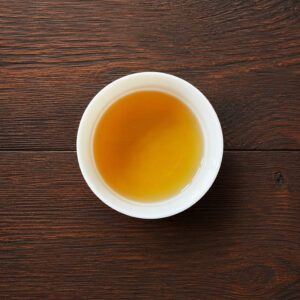 Tasse mit bernsteinfarbenem aufgebrühtem Oolong-Tee auf dunklem Holztisch aus der Draufsicht. Produkt aus dem iūgen Shop.