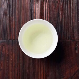 Tasse mit hellgrünem aufgebrühtem Gyokuro-Grüntee auf dunklem Holztisch aus der Draufsicht. Produkt aus dem iūgen Shop.