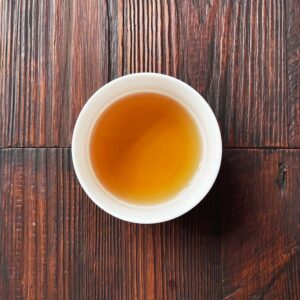 Tasse mit bernsteinfarbenem aufgebrühtem Hōjicha-Tee auf dunklem Holztisch aus der Draufsicht. Produkt aus dem iūgen Shop.