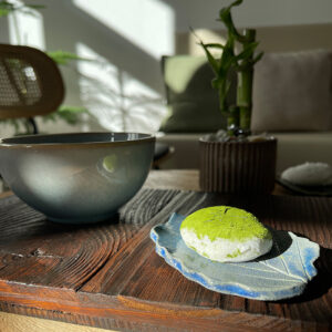 Matcha-bestäubte Süßigkeit auf blauem Keramikteller in Blattform neben einer Schale auf dunklem Holztisch im Sonnenlicht.