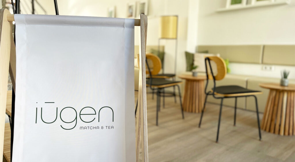 Banner mit dem Logo „iūgen Matcha & Tea“ im Innenraum des Teehauses mit Stühlen und Tischen.
