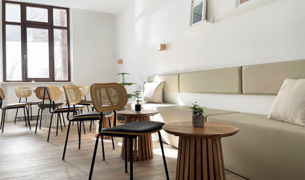 Heller Raum mit Holzstühlen, kleinen runden Tischen und gepolsterter Sitzbank vor großen Fenstern.