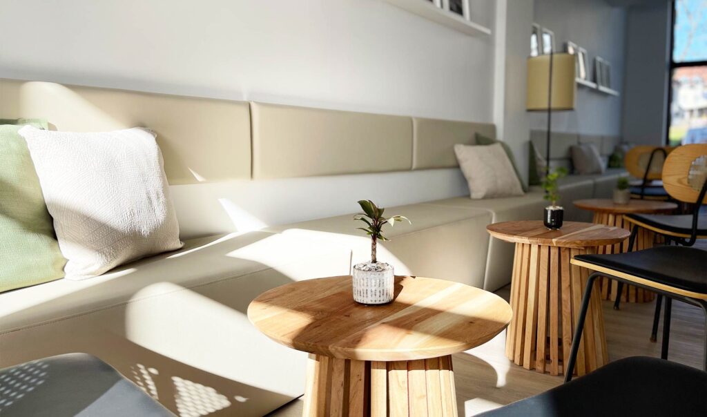 Gemütlicher Sitzbereich bei iūgen mit gepolsterter Bank, kleinen Holztischen und Pflanzen im warmen Tageslicht.