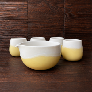 Sonnengelbes Matcha Set mit großer Schale und vier passenden Bechern vor dunklem Holzhintergrund.