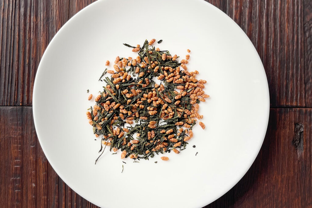 Lose Genmaicha-Teemischung aus grünen Teeblättern und geröstetem Reis auf einem weißen Teller vor dunklem Holzhintergrund. Produkt aus dem iūgen Shop.